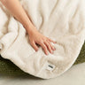 Person holding a fluffy white blanket with a visible brand label on a soft surface.